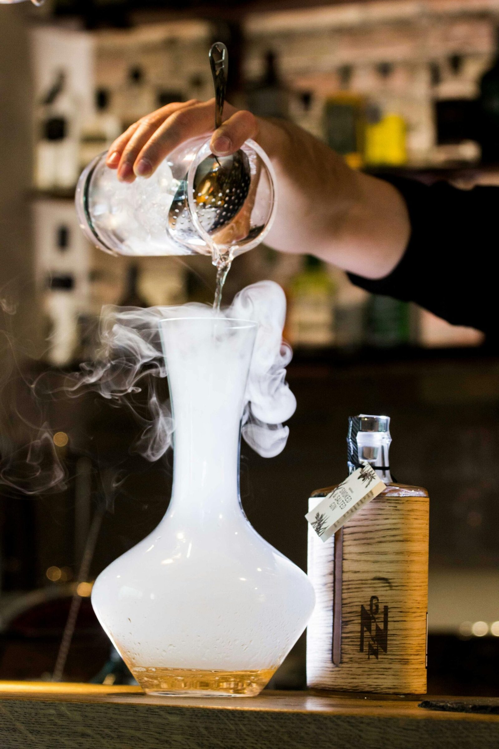 Bartender pouring a cocktail into a misty glass decanter creating a dramatic effect.
